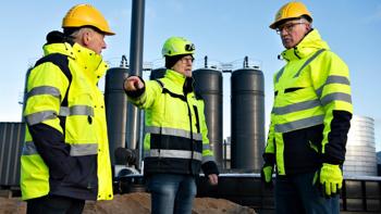 Tre mænd i gult sikkerhedstøj og hjelm står og taler sammen foran biogasanlæg